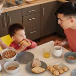 A joyful family of three sharing breakfast in a cozy kitchen setting, featuring waffles and cereal.