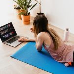 From above side view of young long haired female in casual wear lying on floor on blue mat while using laptop at home